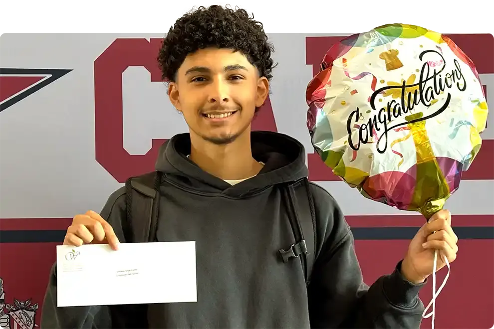 A student smiling as he holds his WAEF award envelope in one hand and a Congratulations balloon in the other, in front of the Eisenhower High School banner
