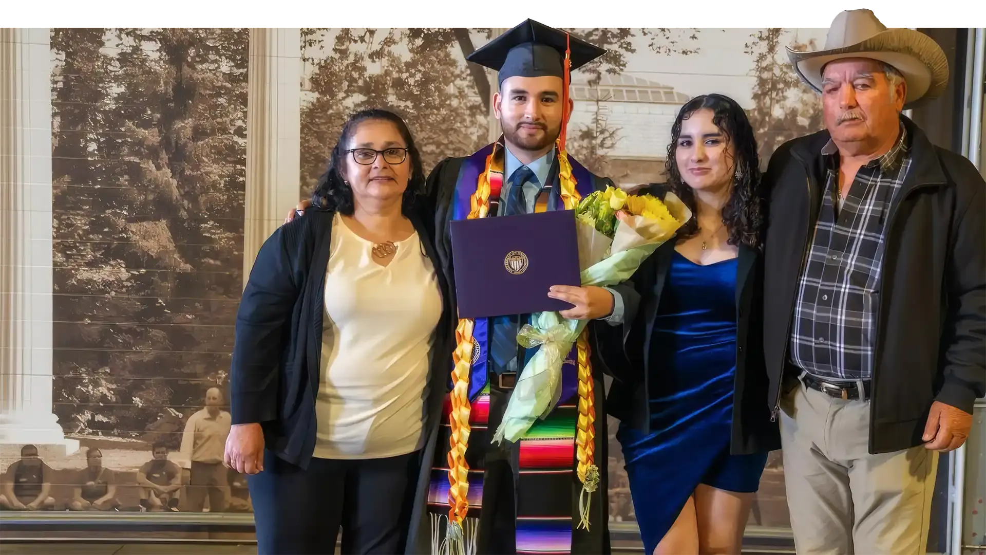 Raul Villanueva in cap and gown with a serape stole, holding his diploma and a bouquet, his mother, sister and father beside him at his graduation