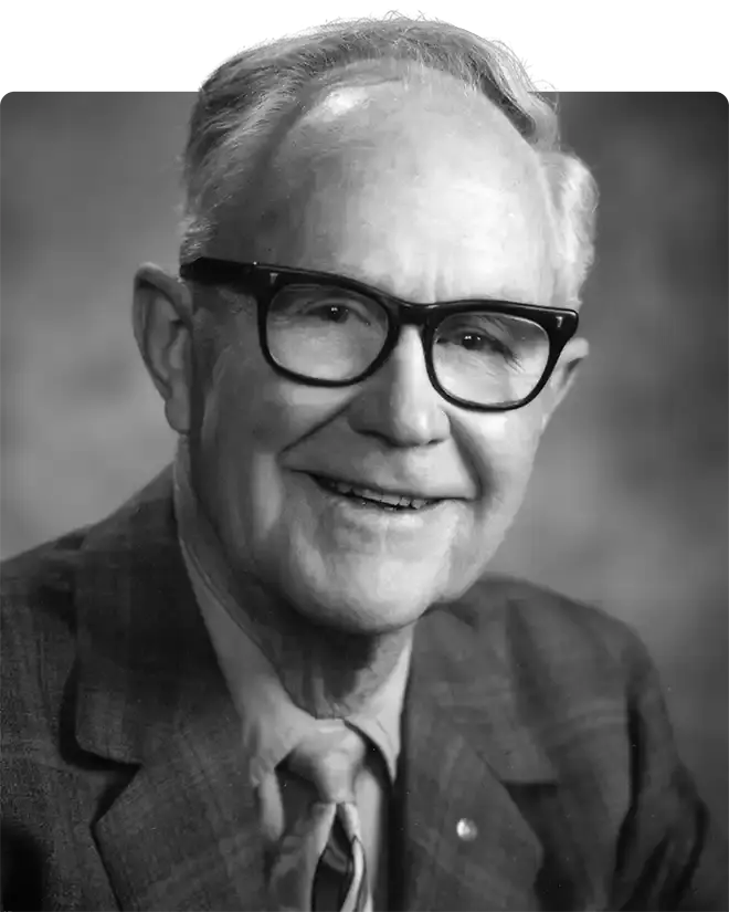 William "Bill" Luce, in a black and white portrait, smiling in heavy-rimmed glasses, a checked jacket and a tie