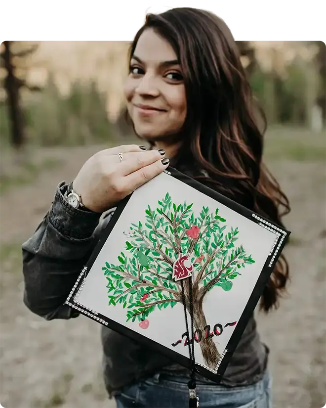 Dianna Sanchez smiling over her shoulder and holding up her mortarboard, hand-painted with an apple tree in fruit and lettered 2020