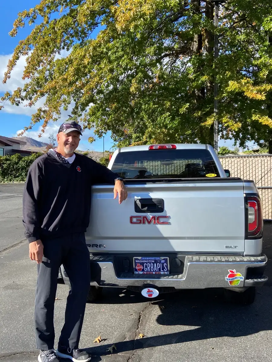 A man in a navy pullover and cap leaning on the tailgate of a silver GMC Sierra with a personalized Washington Apples plate reading GR8APLS