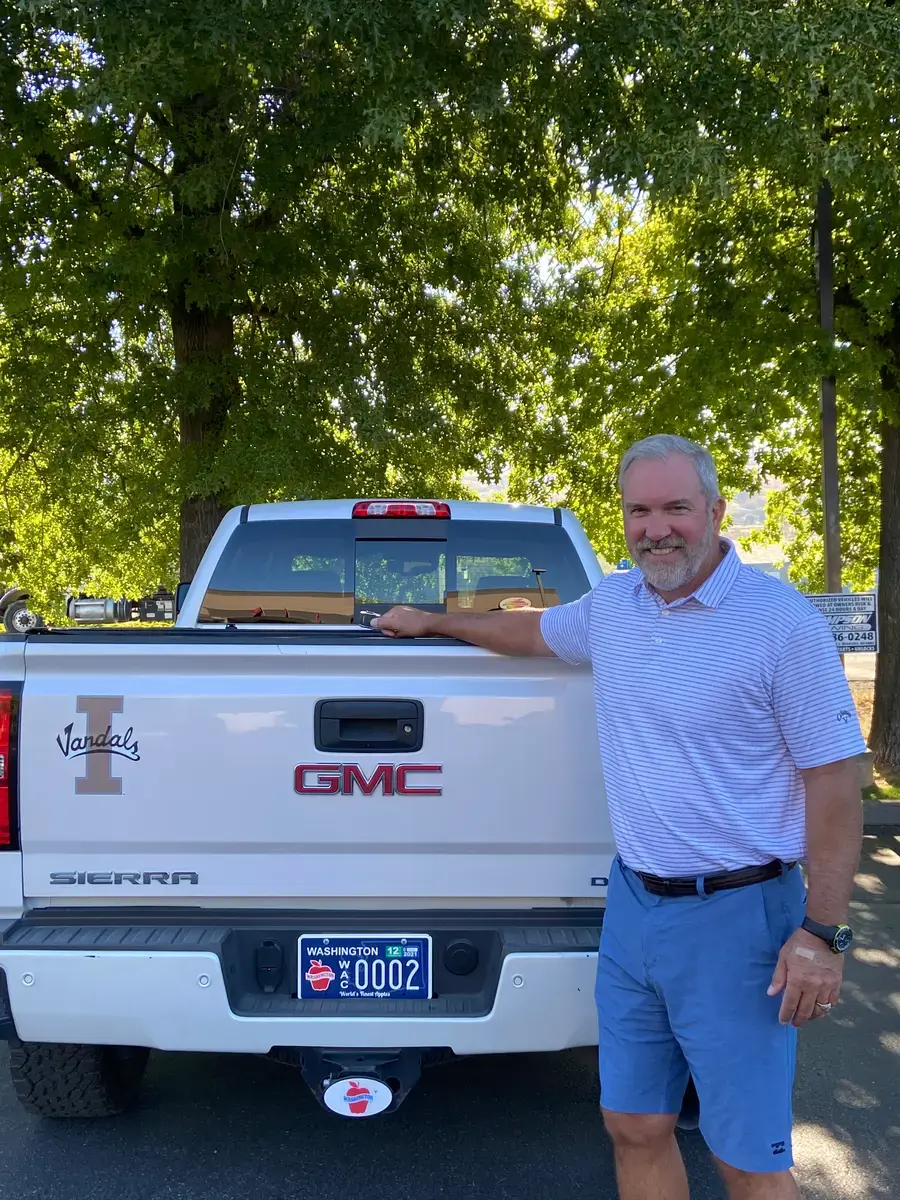 A man in a light blue polo beside the bed of a white GMC Sierra with a Washington Apples plate, parked under green trees