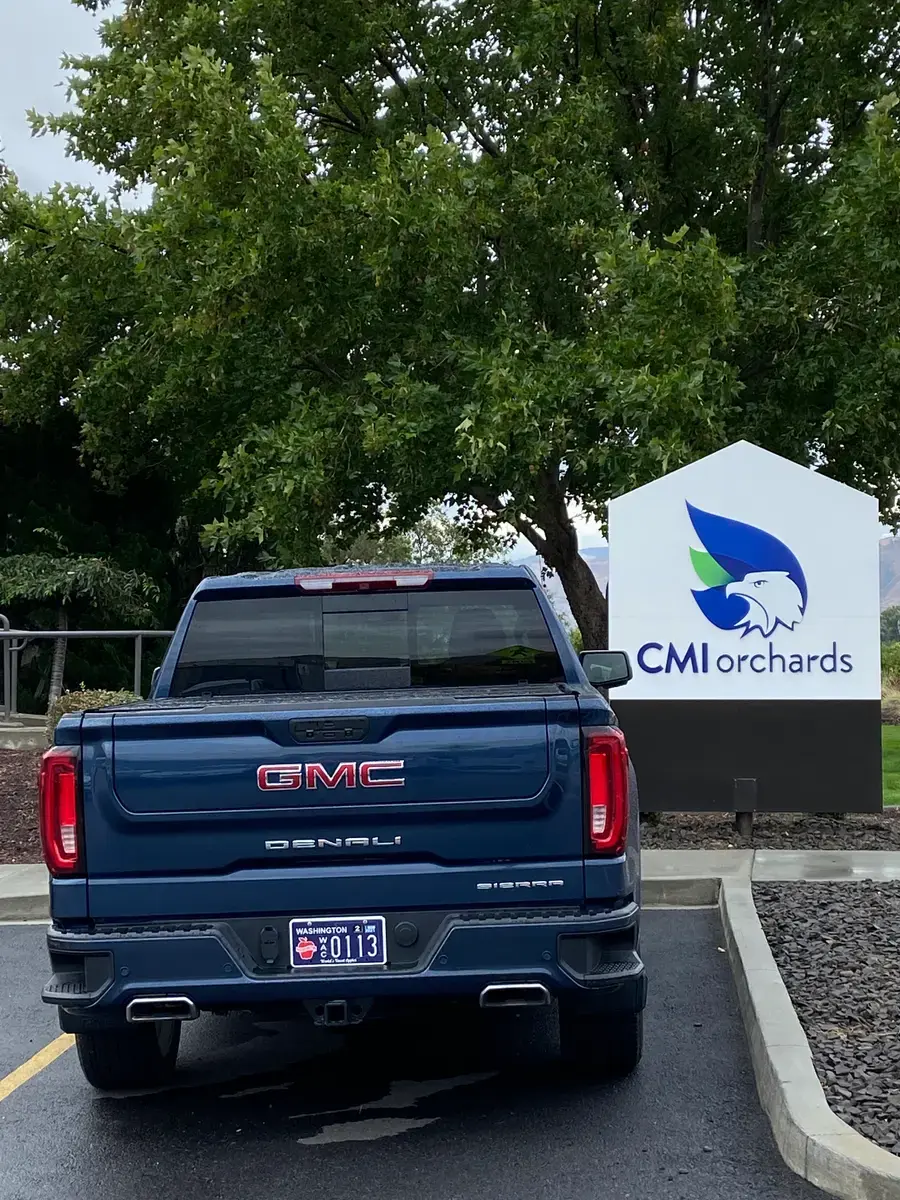 A dark blue GMC Denali with a Washington Apples plate, parked at the CMI Orchards sign