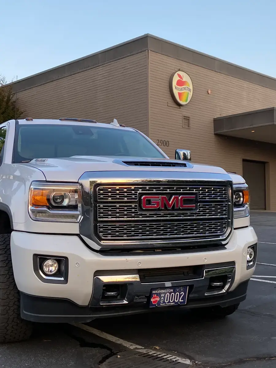 The front of a white GMC Denali with a Washington Apples plate, parked outside the Washington Apple Commission building