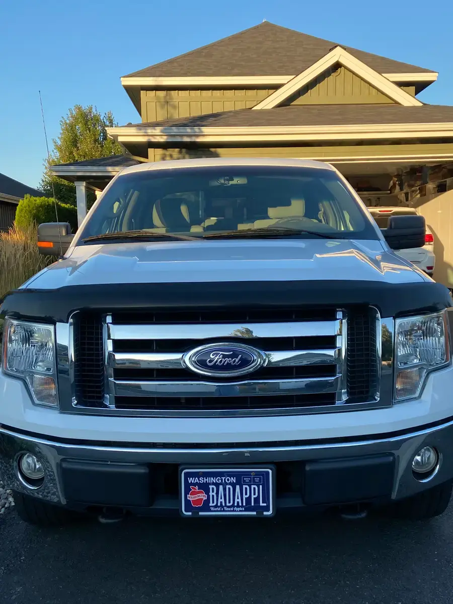 The front of a white Ford F-150 in a driveway with a personalized Washington Apples plate reading BADAPPL