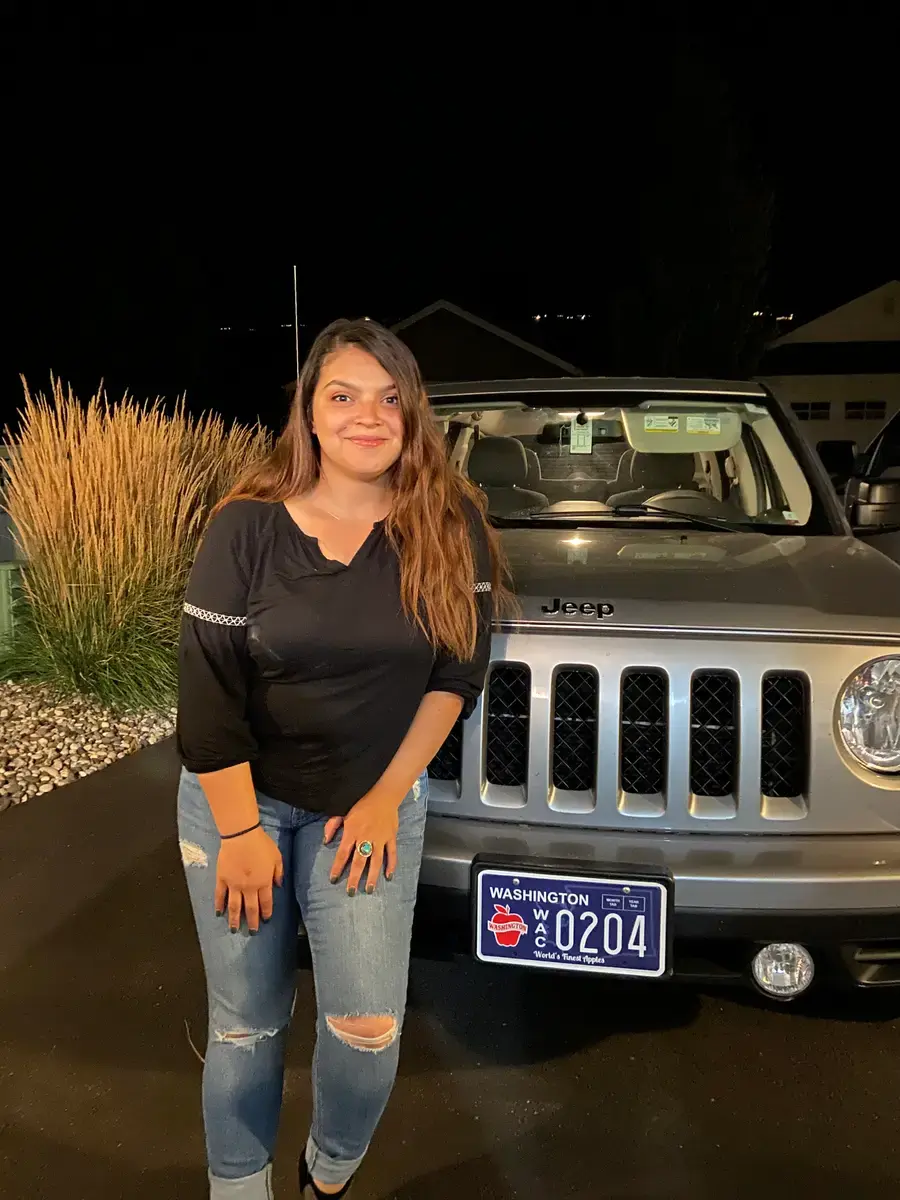A woman standing at night beside a silver Jeep Patriot with a Washington Apples plate, lit by a porch light