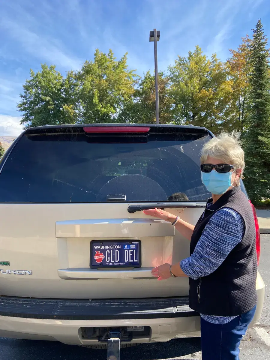 A woman in a face mask pointing at the personalized Washington Apples plate on the back of a tan SUV, reading GLD DEL