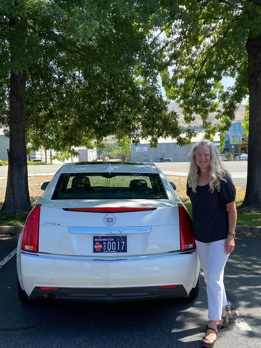 A woman with long fair hair standing beside the back of a white Cadillac sedan with a Washington Apples plate, under a shade tree