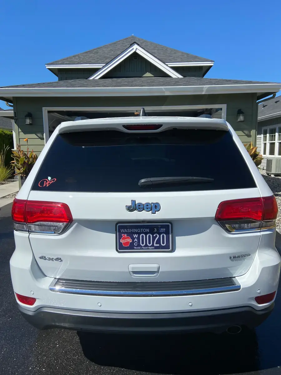 The back of a white Jeep Grand Cherokee with a Washington Apples plate, in the driveway of a grey house