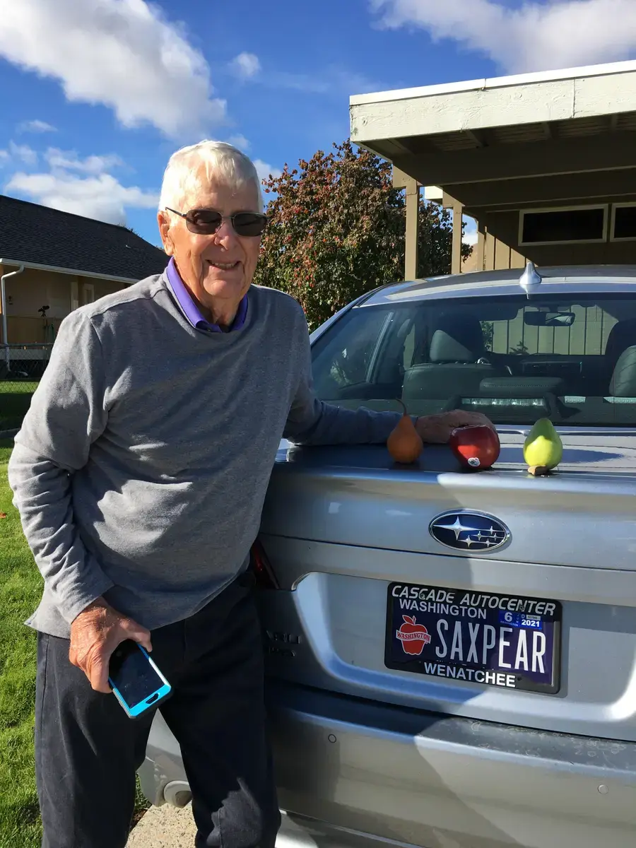 A man in sunglasses beside a silver Subaru with a personalized Washington Apples plate reading SAXPEAR, an apple and a pear set on the tailgate