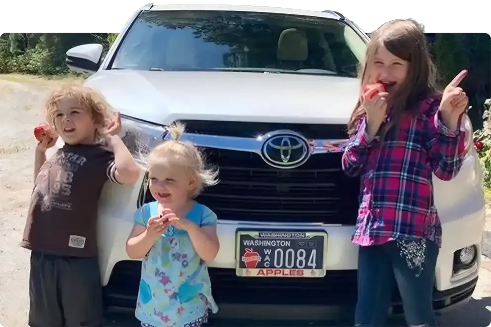 Three young children eating apples in front of a white SUV fitted with a Washington Apples specialty plate