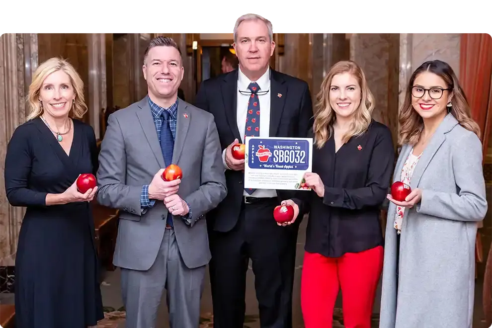 Five supporters holding red apples and a mock Washington Apples plate reading SB6032, in a marble hallway in Olympia