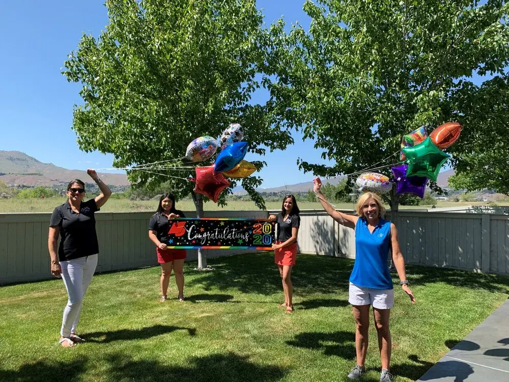 Four people celebrating on a lawn beside a congratulations sign and balloons