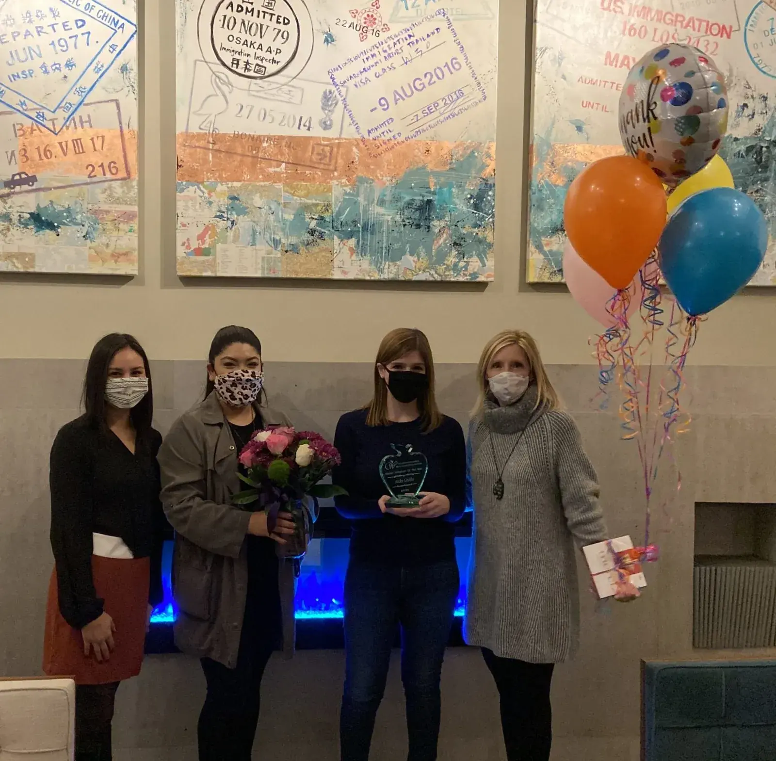 Four people in face masks standing together, one holding flowers and an award, balloons alongside