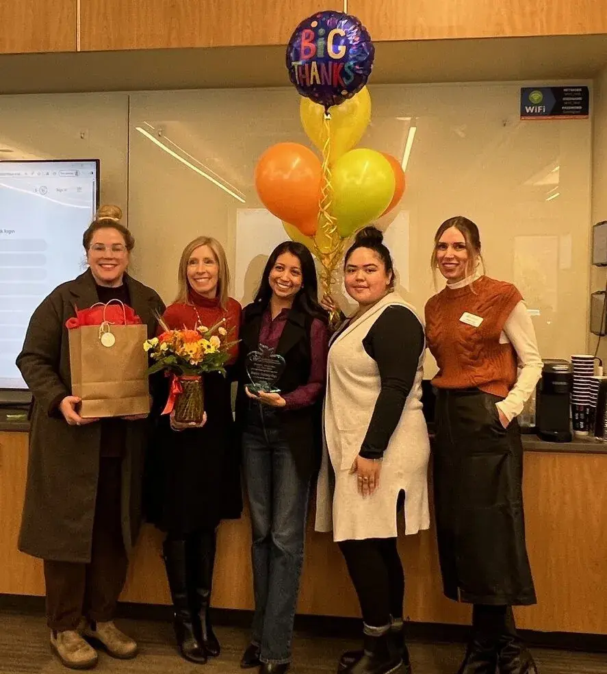 Five women standing together with flowers, a gift bag and an award, balloons overhead