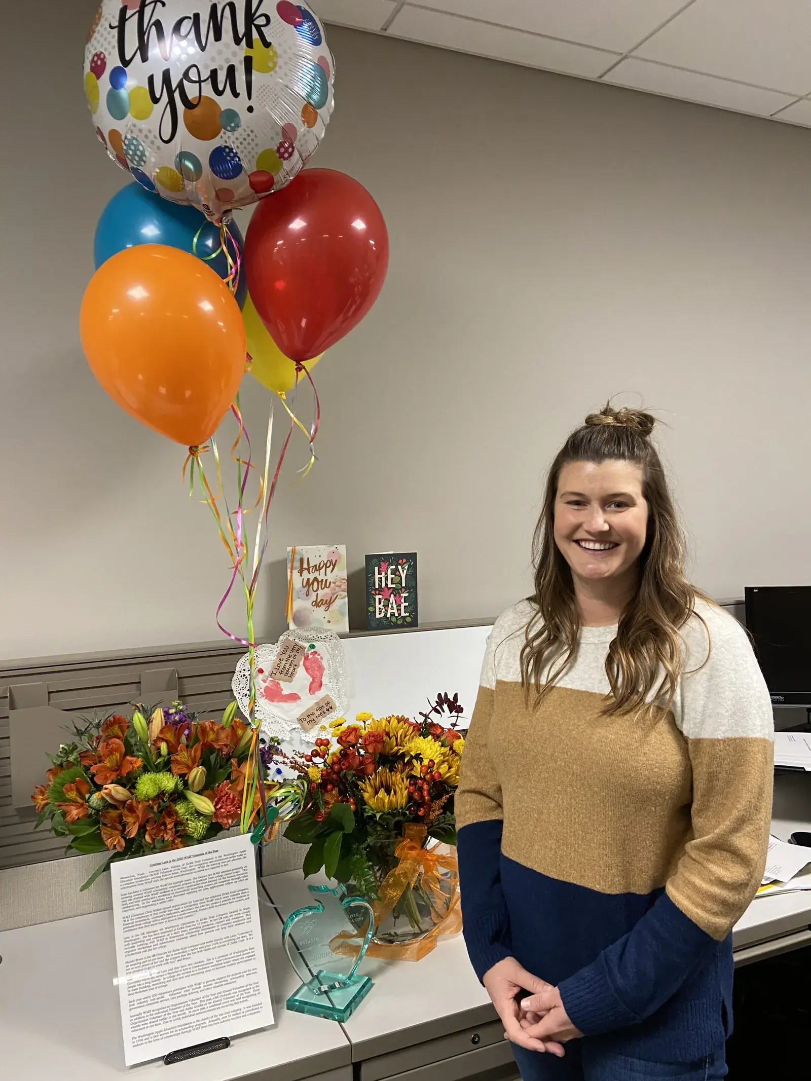 A woman standing at a desk with flowers and a bouquet of thank-you balloons