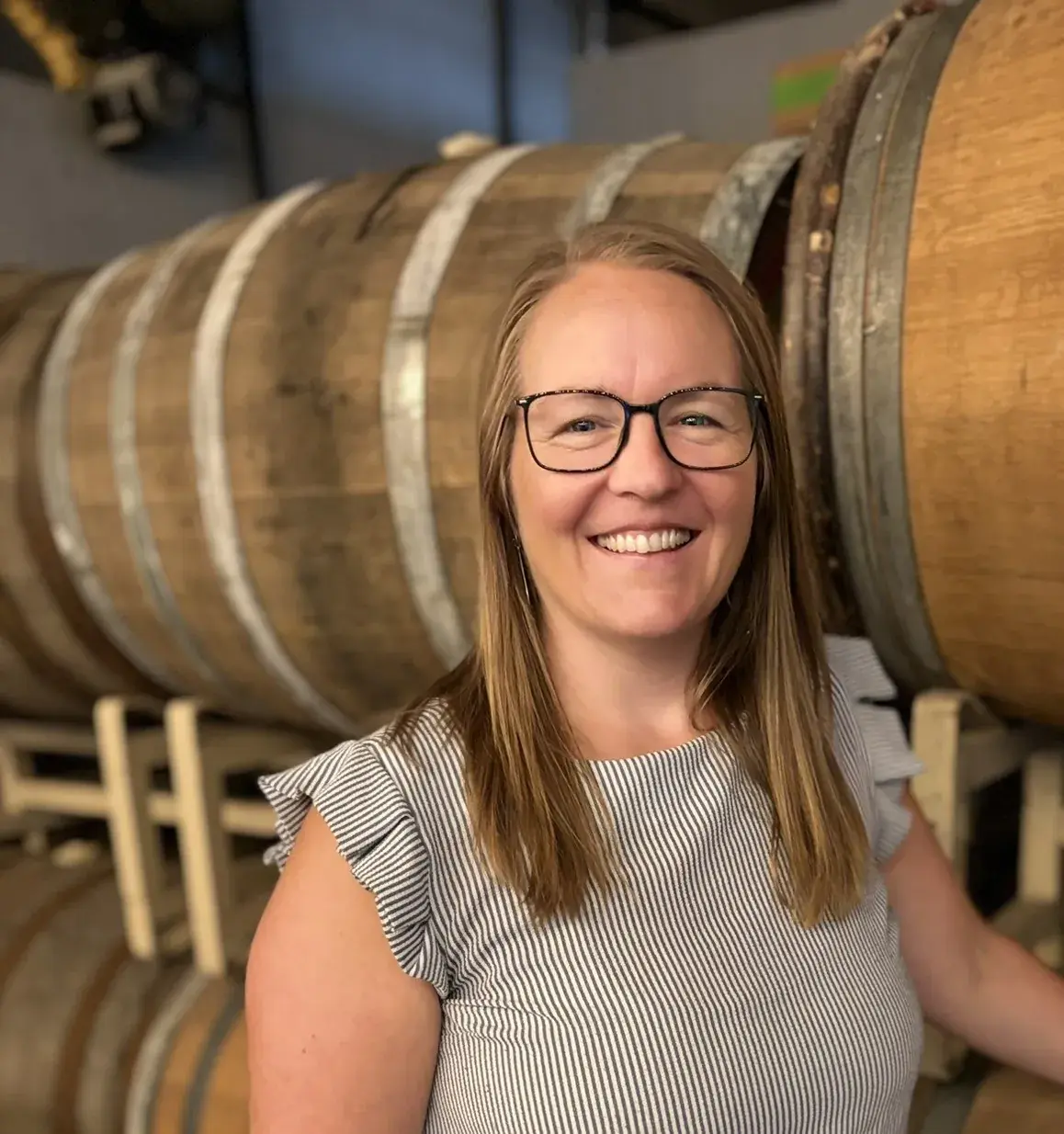 A woman in glasses smiling beside a stack of wooden barrels