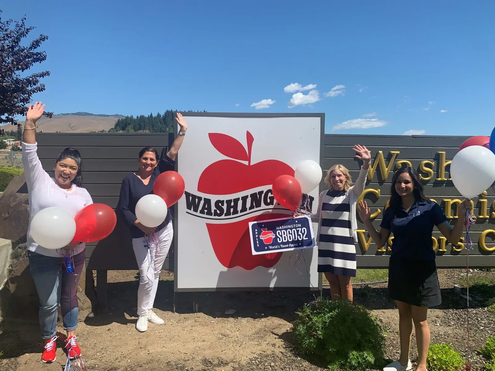 Four people with balloons beside a large Washington Apples licence plate sign