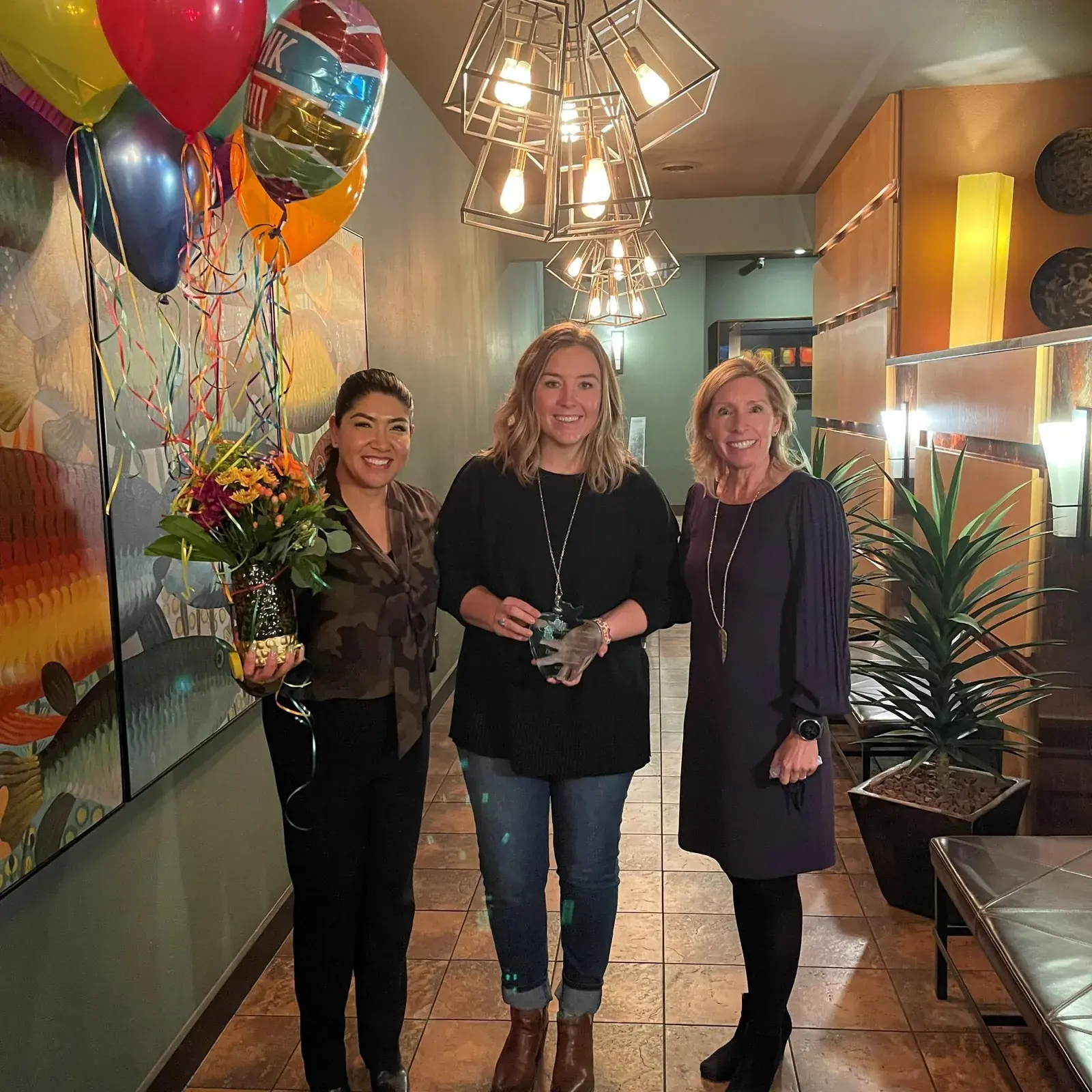 Three women standing together, one holding a potted plant, with balloons behind them