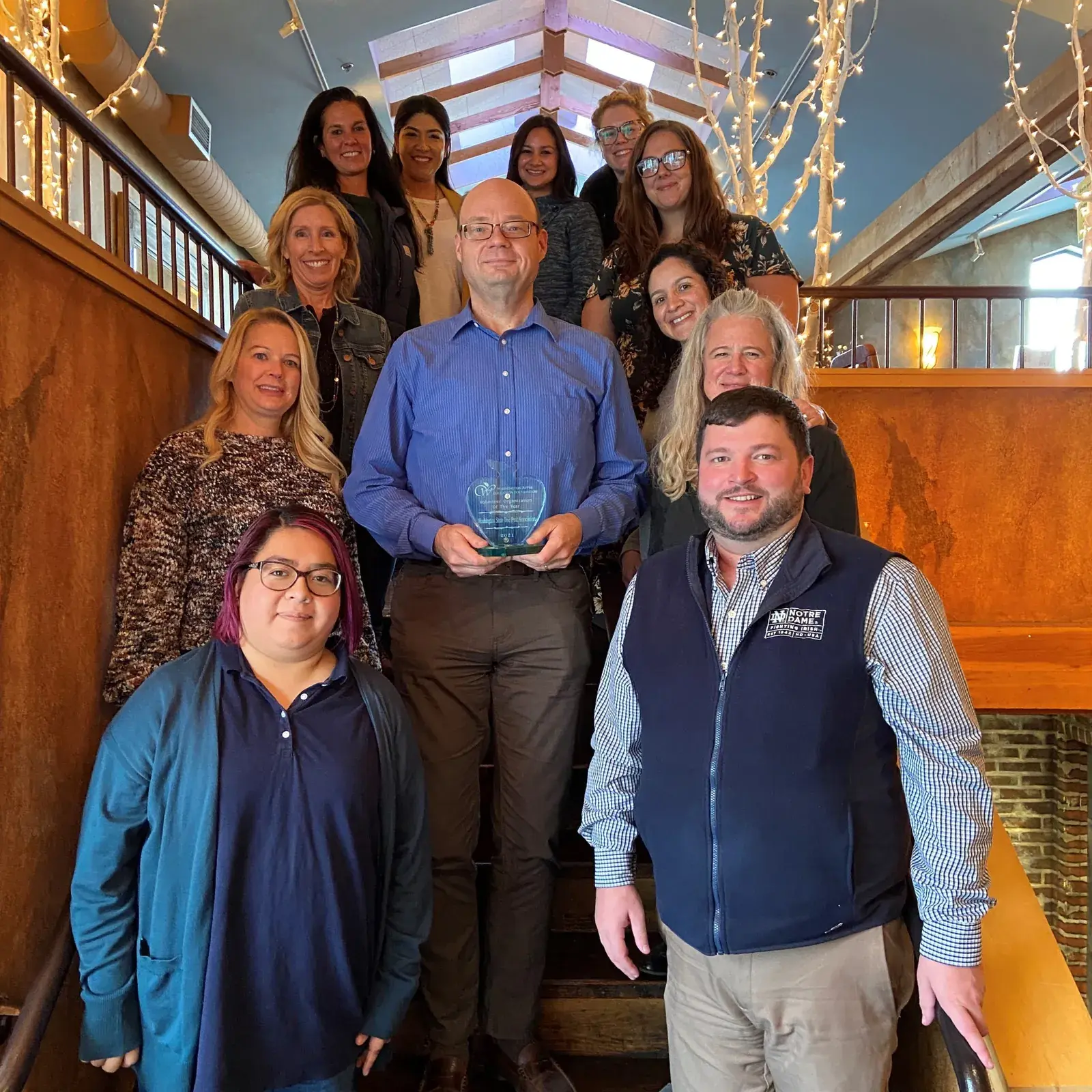 A large group posed on a staircase, one man holding a glass award