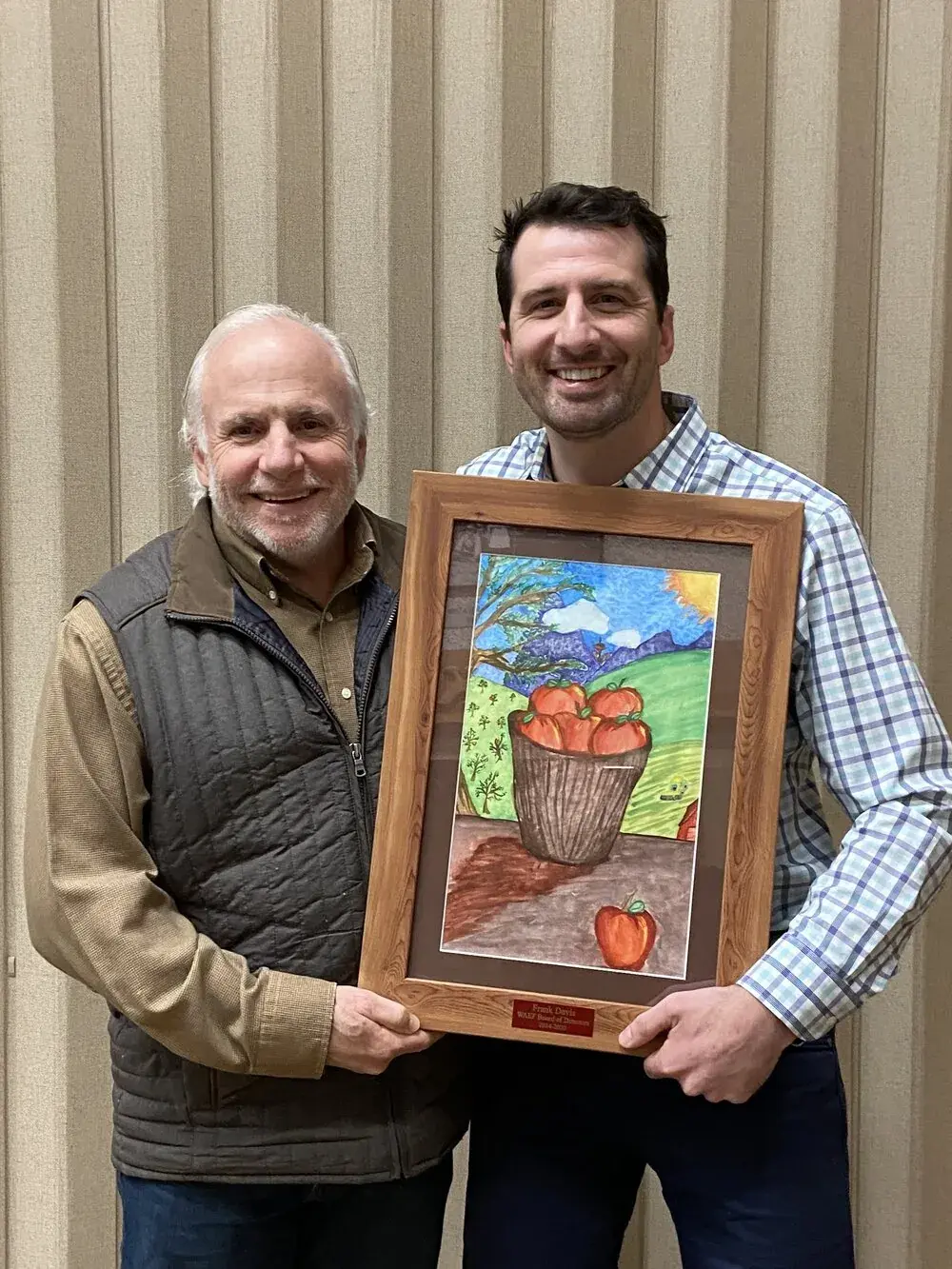 Two men holding a framed painting of a basket of apples