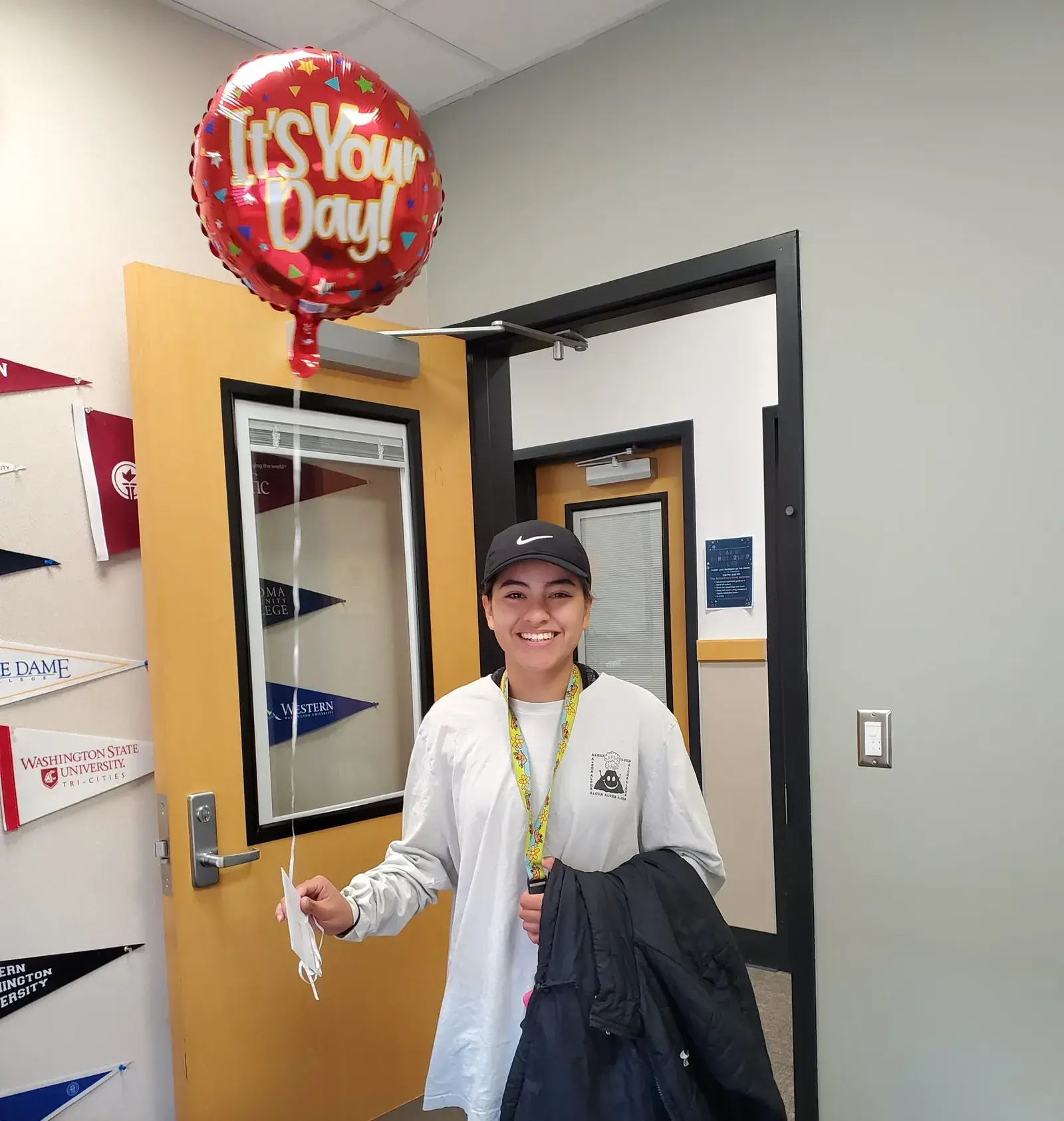 A student in a school hallway holding a pen, an It's Your Day balloon overhead