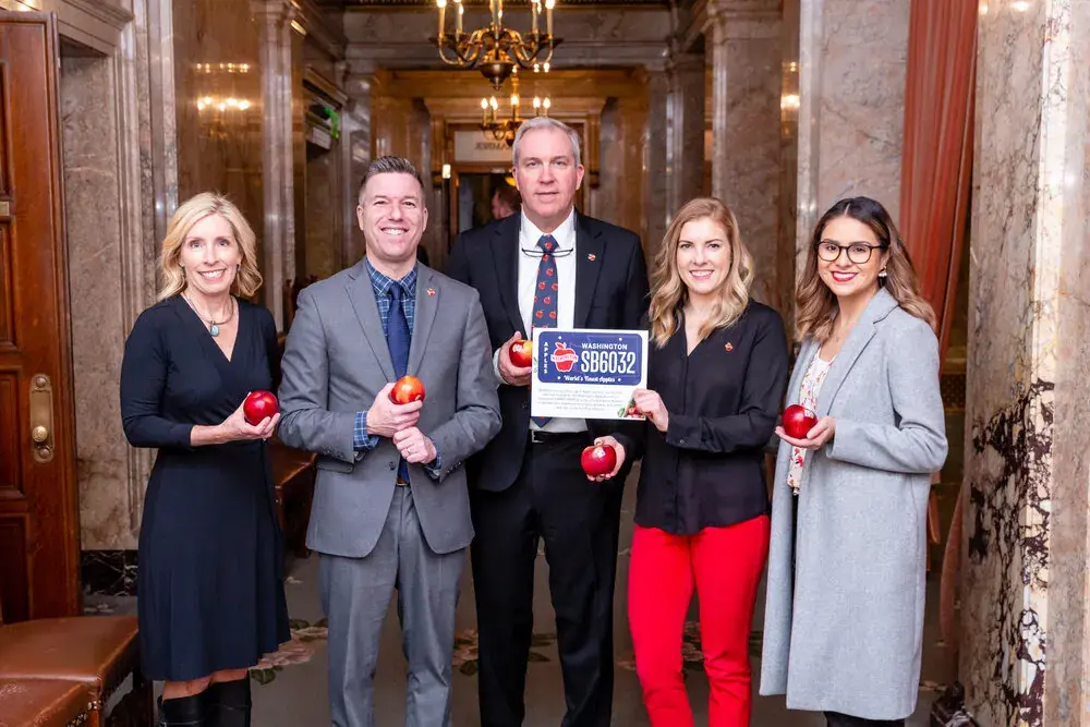 Five people holding apples and a sign inside a state capitol building