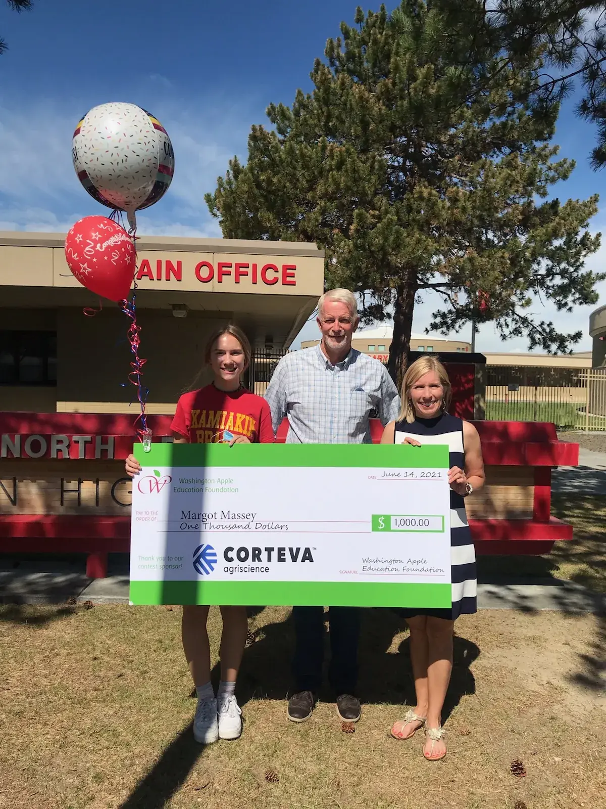 Four people holding an oversized Corteva cheque outside a school, balloons overhead