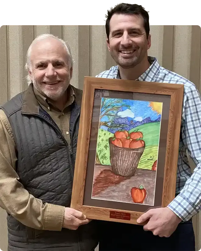 Two WAEF board members smiling as they hold up a framed student painting of a basket of apples