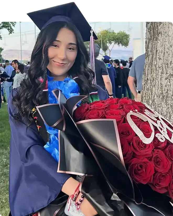A smiling graduate in a navy cap and gown and a blue lei, holding a large bouquet of red roses at commencement