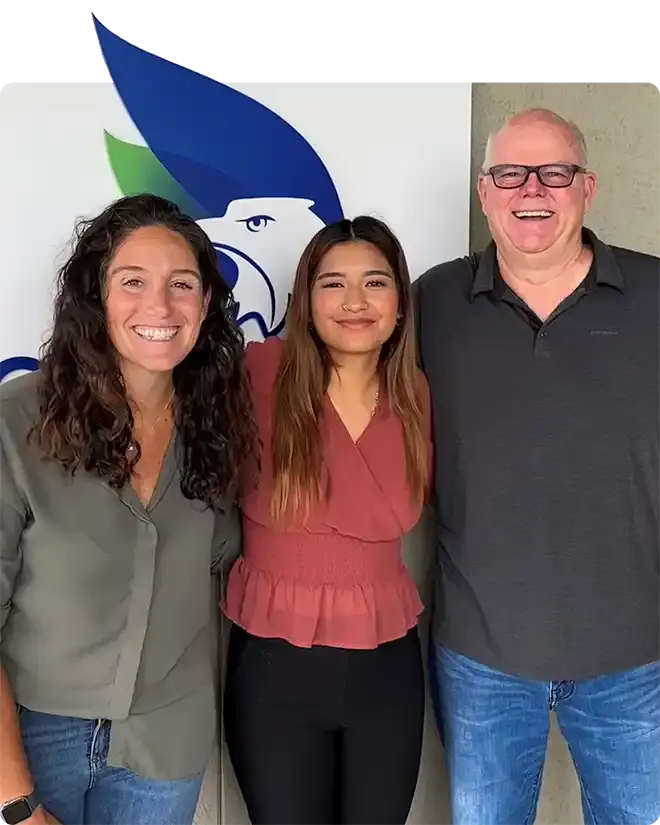 A smiling student in a rose blouse standing between a woman and a man in glasses, in front of a blue and green eagle logo