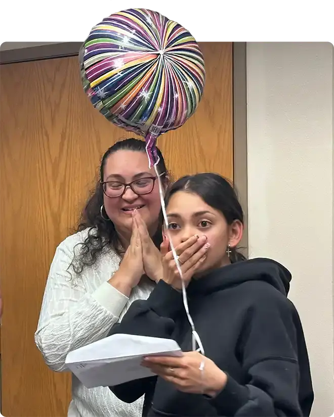 A student with her hand over her mouth in surprise, holding her award letter, her mother beside her with hands clasped and smiling, under a foil congratulations balloon