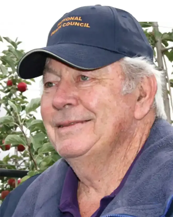 Fred Valentine, smiling, in a navy National Peach Council cap, in front of apple trees heavy with red fruit