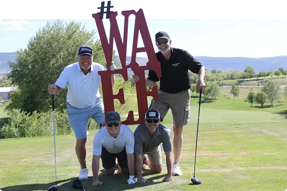 Four golfers posing with a large red WAEF sign on the green at a WAEF golf tournament, two kneeling in front and two standing with their drivers
