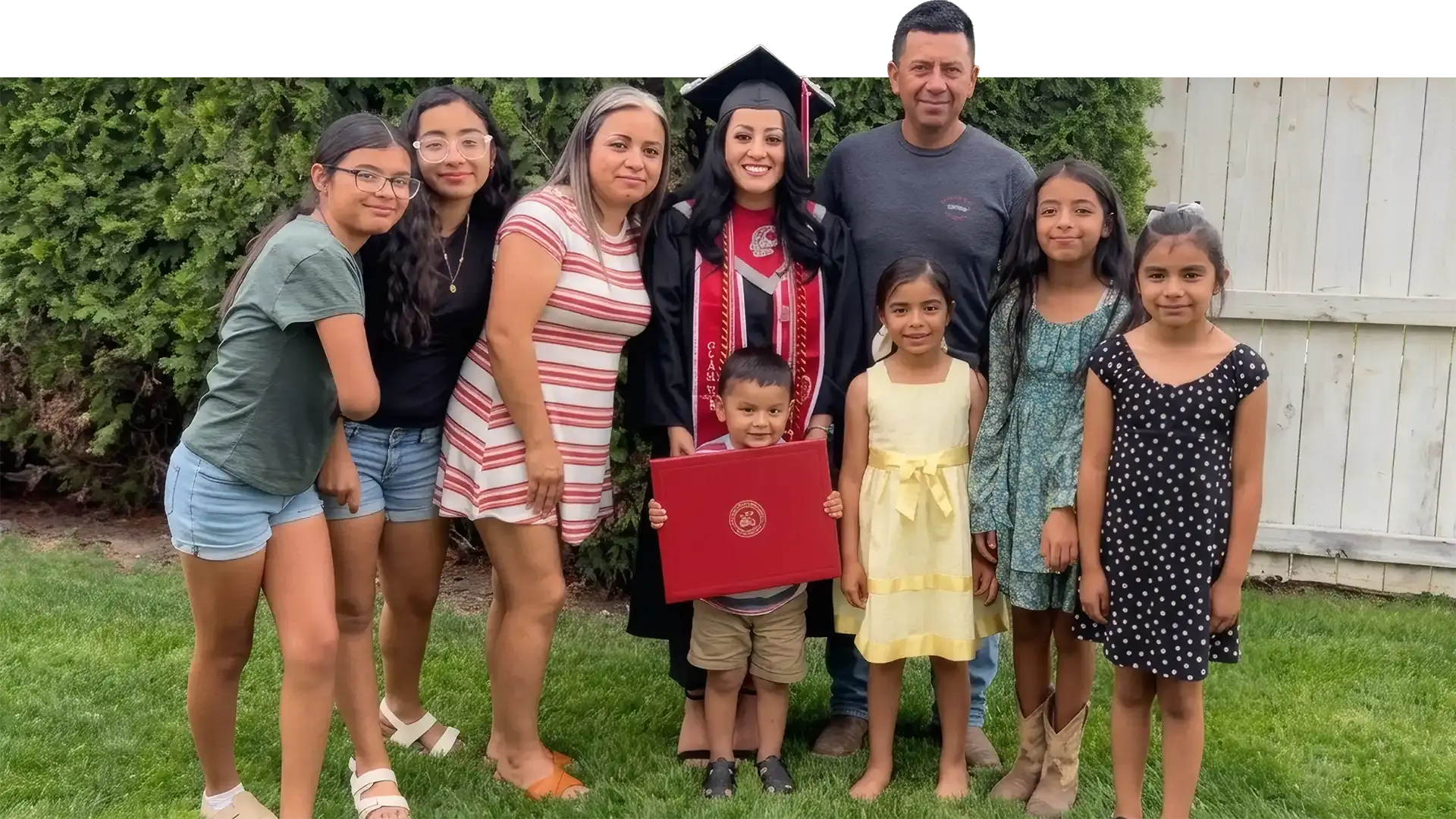 A WAEF graduate in cap, gown and stole holding her diploma on the lawn at home, her parents, sisters, cousins and a small boy gathered around her