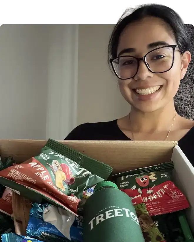 A WAEF student in glasses smiling over an open care package box of apple chips, snacks and a Tree Top water bottle
