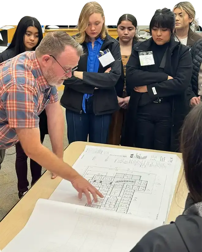 WAEF students gathered around a table while a man in a plaid shirt points out a detail on a large architectural floor plan
