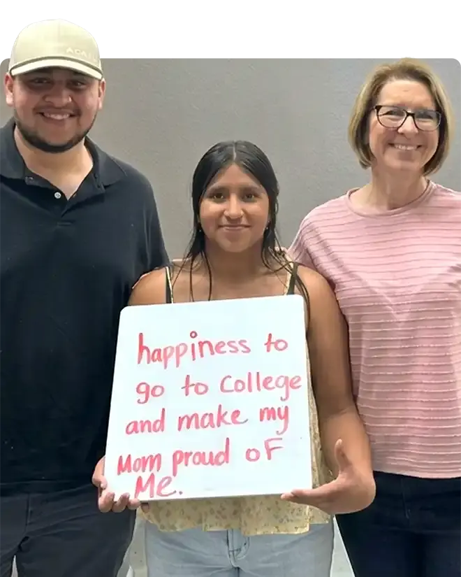 A WAEF scholarship recipient standing between two adults, holding a whiteboard she has written on in red — "happiness to go to college and make my mom proud of me"