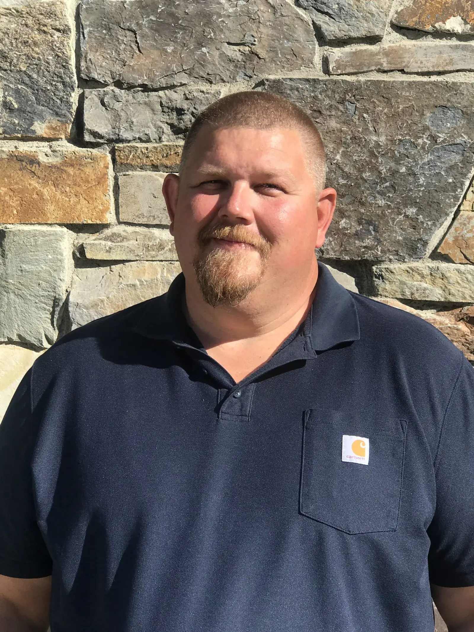Keith Veselka, in a navy work shirt, standing in front of a stone wall