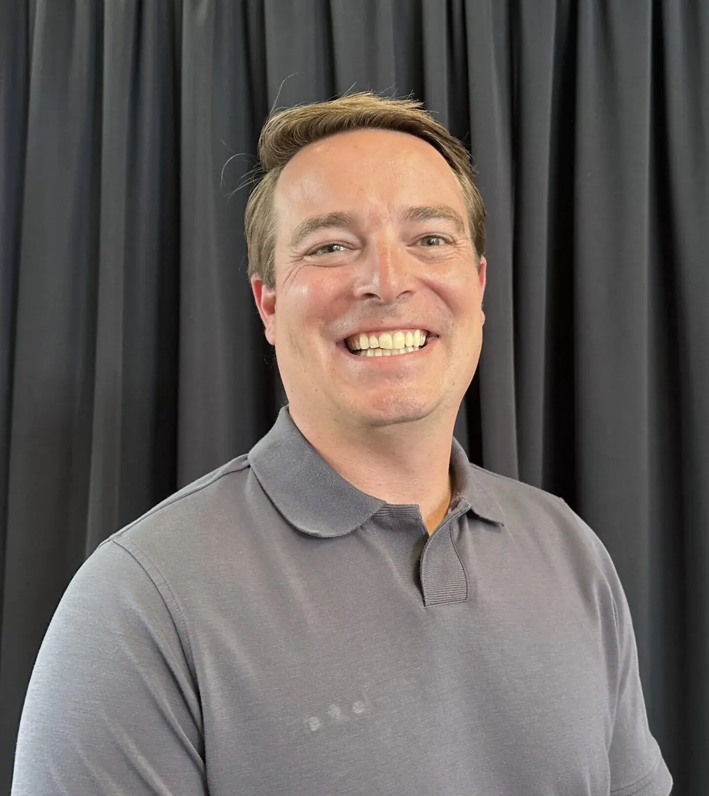 Brandon Flory, in a grey polo shirt, in a studio portrait against a dark curtain