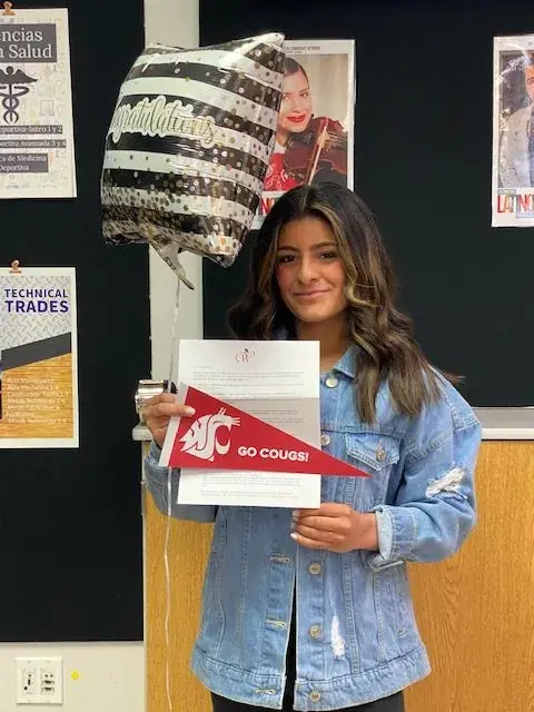 A student in a denim jacket holding a Washington State University pennant and an envelope, a balloon overhead