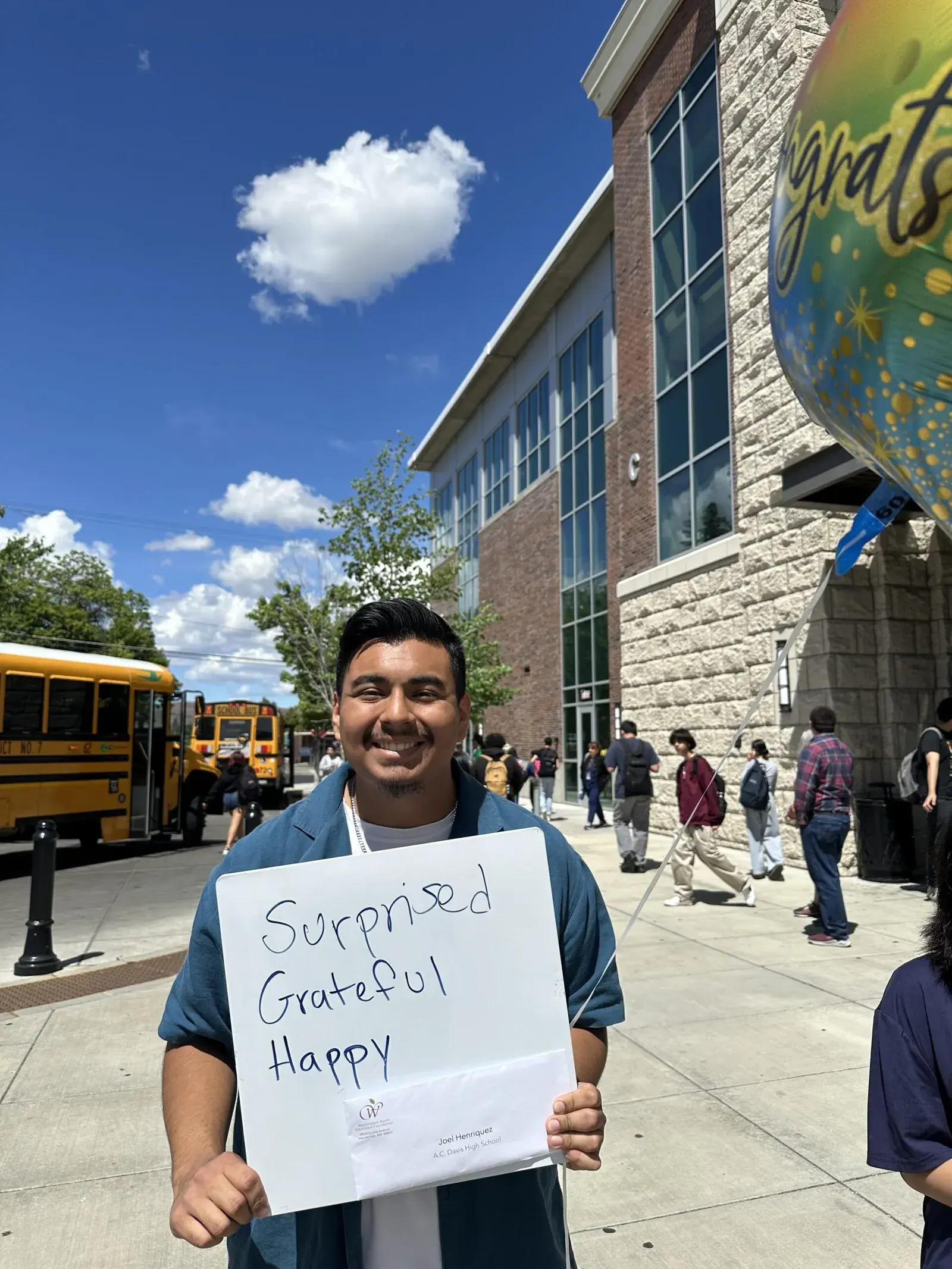 A student outdoors holding a handwritten sign reading Surprised, Grateful, Happy, with school buses behind him