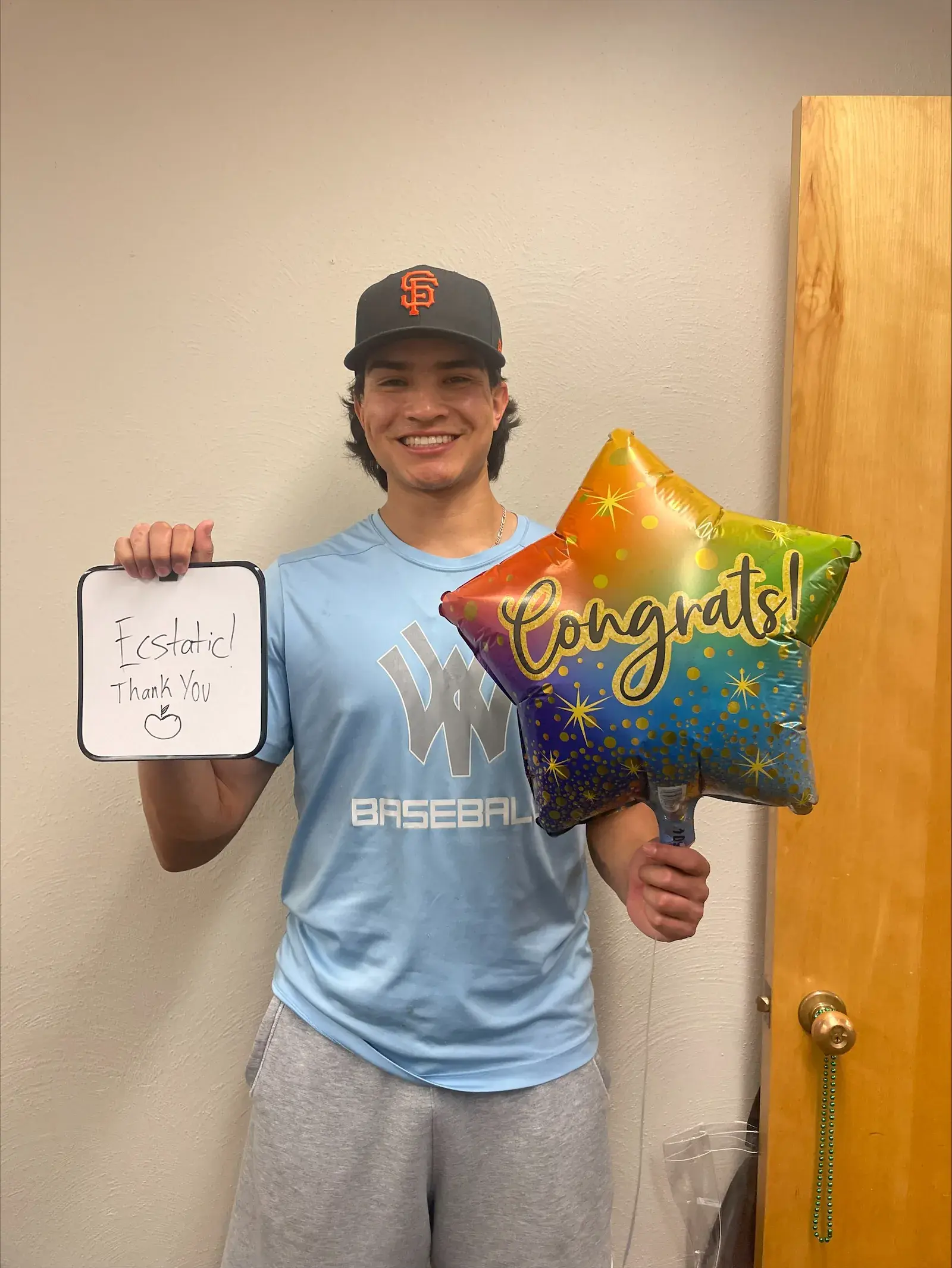 A student in a baseball cap holding a small whiteboard and a congratulations balloon