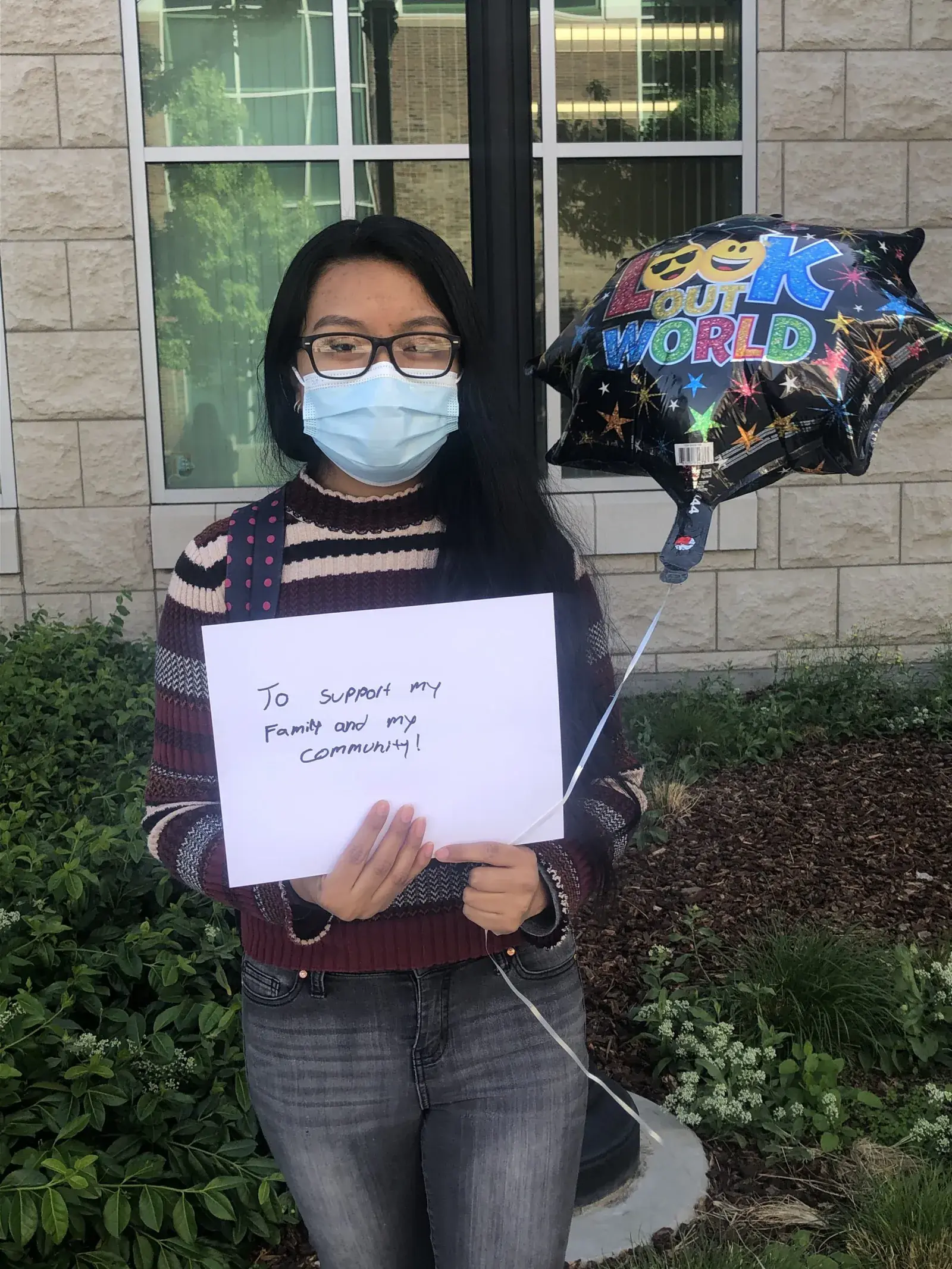 A student in a face mask standing outdoors holding a handwritten thank-you note and balloons