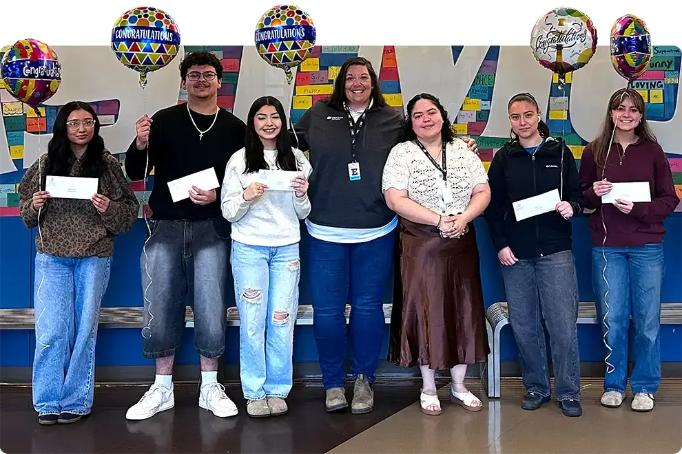 Eight scholarship recipients holding their award envelopes, with congratulations balloons, lined up in front of the colorful I Am Eastmont wall in the school commons