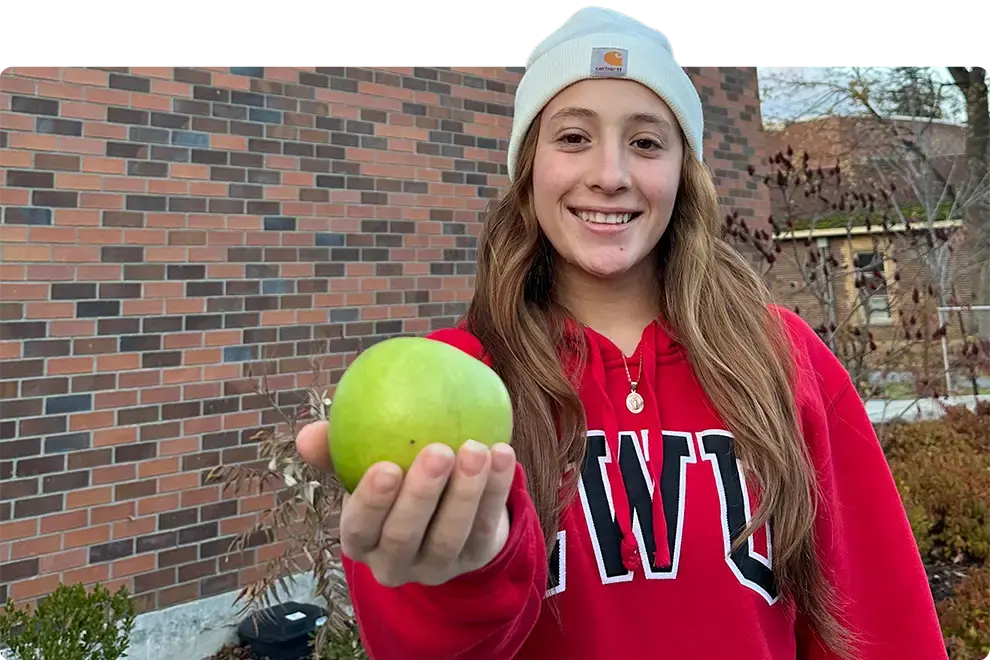 A WAEF student in an EWU sweatshirt holding out a green apple outside a brick campus building
